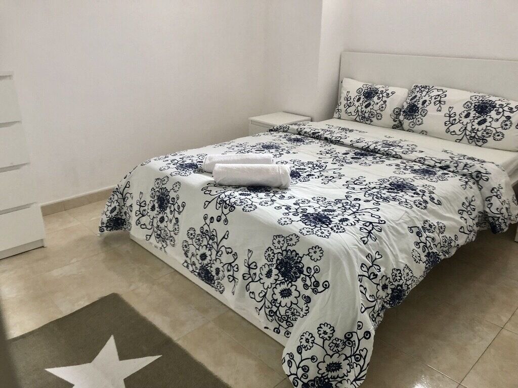 A neatly made bed with a floral-patterned bedspread and matching pillows, a bedside table, and a star-patterned rug on a tiled floor.