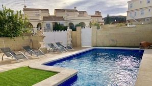 Una piscina rettangolare con bordo piastrellato, circondata da un basso muro e da una zona erbosa. Si vedono lettini e un cancello bianco sullo sfondo.