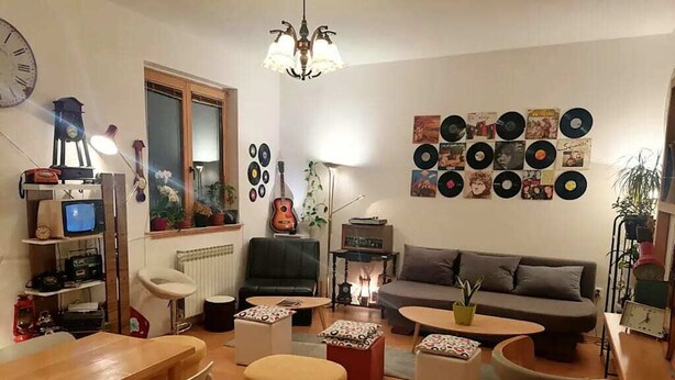 Una sala de estar acogedora con un sofá, una mesa de centro y otomanas. Se aprecia una pared con discos de vinilo, un reloj y una lámpara.
