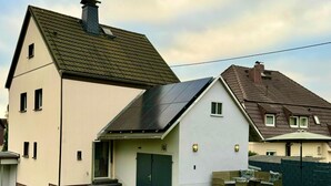 Ett tvåvåningshus med en solpanel på taket, en grön dörr och en liten veranda med utemöbler.