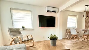 Ett modernt vardagsrum med trägolv, en grå soffa, trästolar, ett soffbord, en TV som är monterad på väggen och en krukväxt.