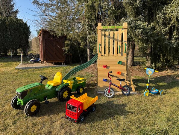 Ein Spielplatz mit einer hölzernen Kletterkonstruktion, einer Rutsche und verschiedenen Spielgeräten, darunter ein Dreirad und Spielzeugautos.