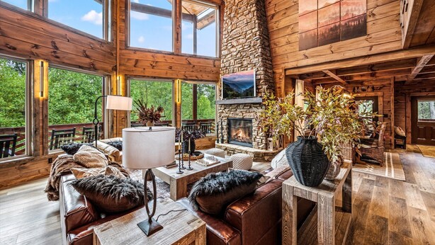 Un intérieur spacieux de chalet en bois, avec une cheminée en pierre, des meubles en bois et de grandes fenêtres offrant une vue sur l’extérieur.