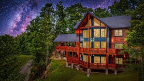 Une grande cabane en rondins à plusieurs étages, dotée d’un balcon imposant, entourée d’une végétation luxuriante et surplombant un ciel nocturne étoilé.