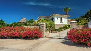 A villa with a stone entrance, lush pink flowering plants, and a thatched-roof structure in the background.