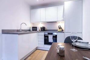 A modern kitchen with white cabinets, a microwave, a coffee maker, and a dining table set with a wine glass and plates. - Trendy Apt in the Heart of MCR (Manchester)