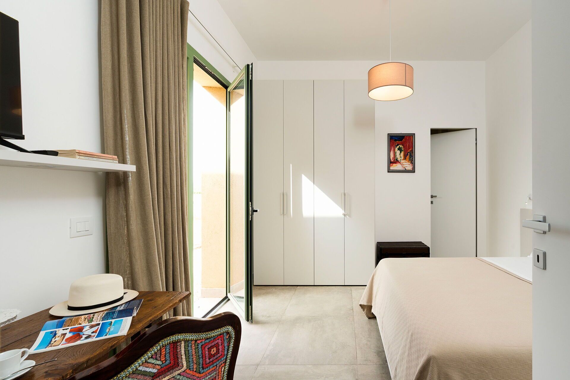 Une chambre d’hôtel avec un lit, un bureau sur lequel se trouvent un chapeau et un magazine, un téléviseur fixé au mur et un tableau accroché au mur.