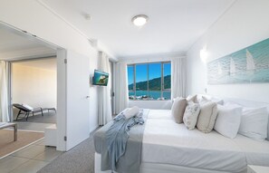 A bedroom with a bed, a TV, a window with a view of the sea, and a painting on the wall.