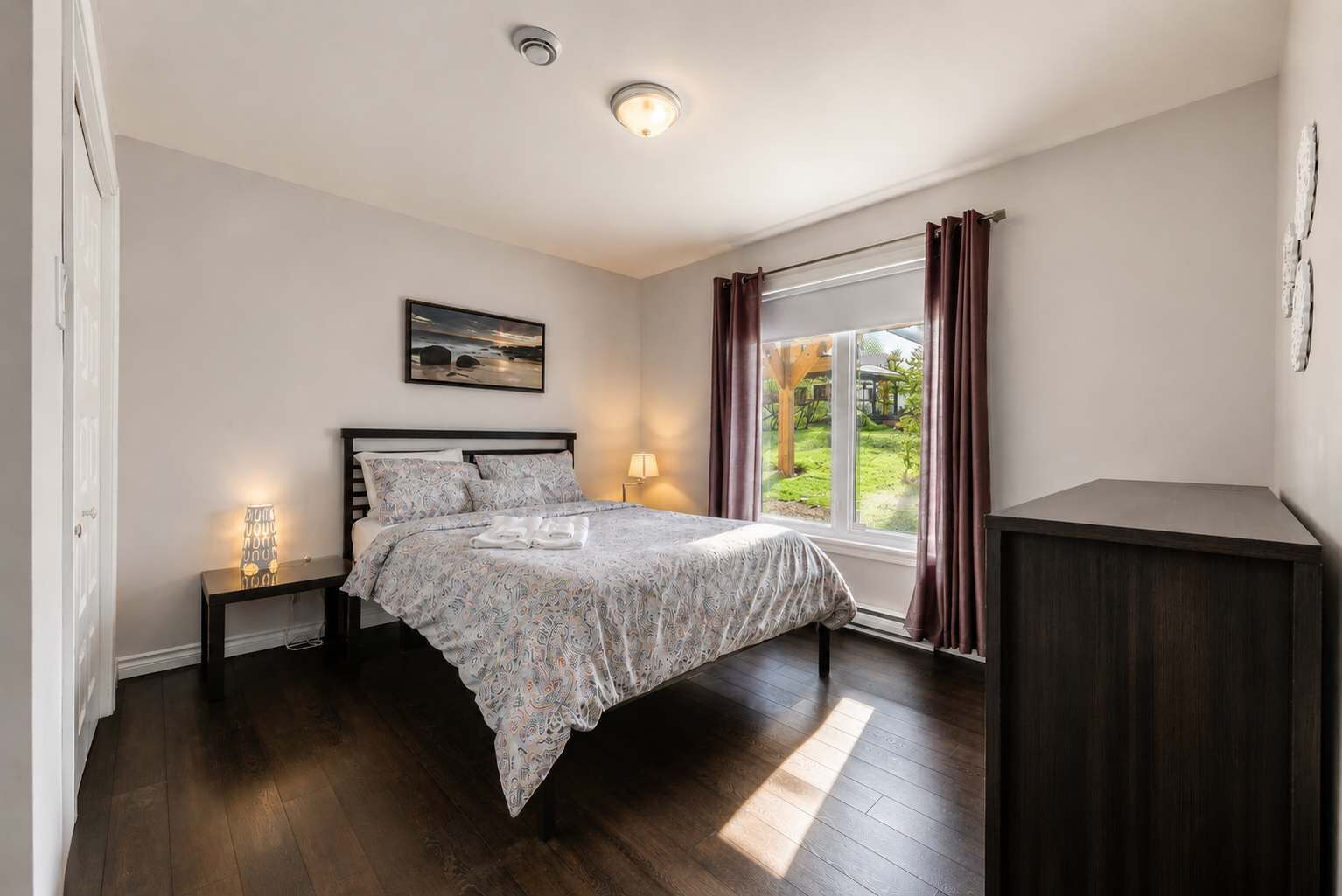 A bedroom with a bed, bedside lamps, a window with curtains, a view of greenery, and a small cabinet.