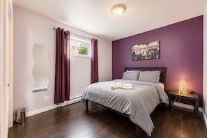 A bedroom with a bed, a nightstand, a window with curtains, a wall-mounted mirror, and a cityscape painting.