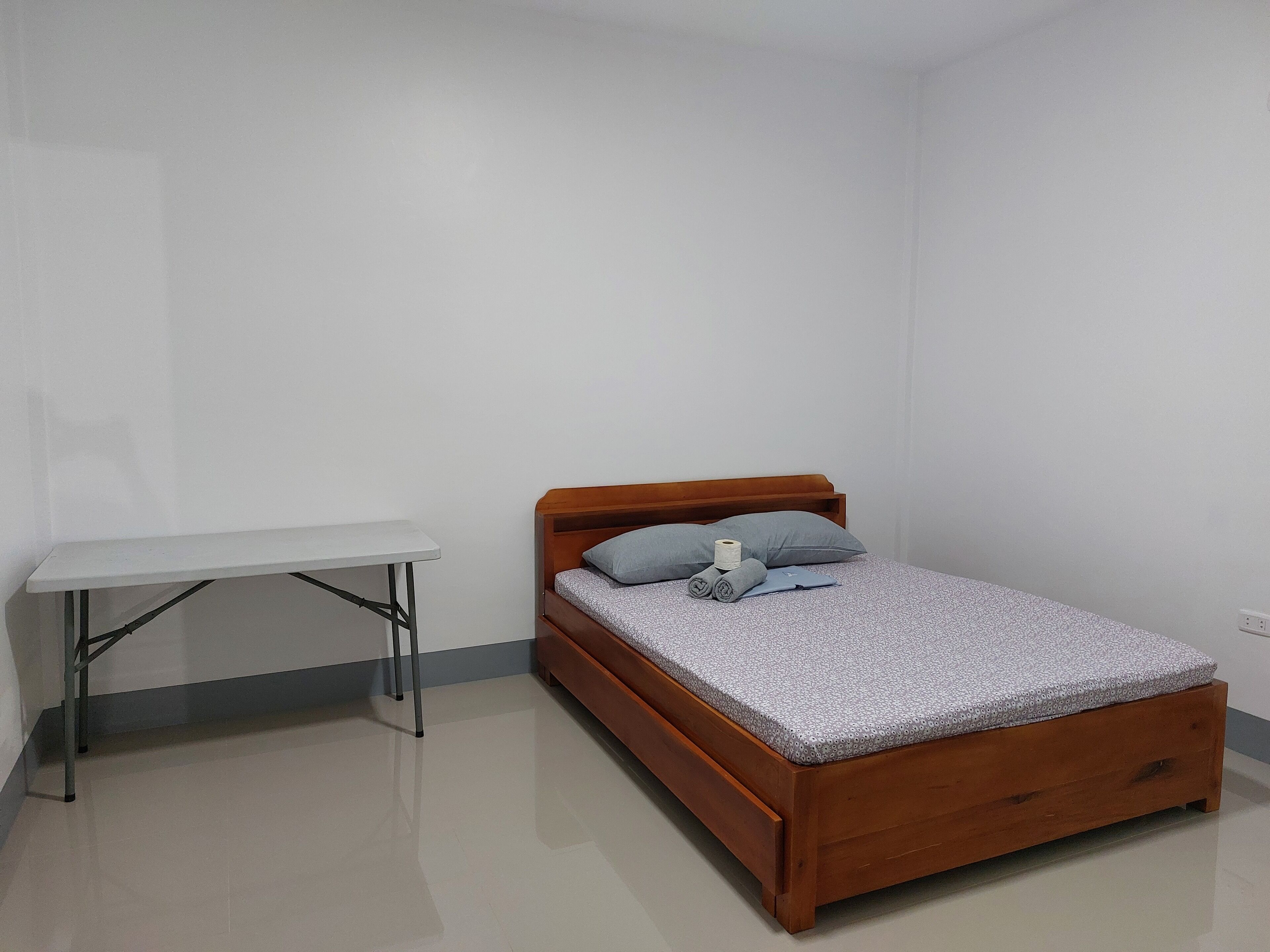 Quarto pequeno e bem organizado, com uma cama de madeira, uma escrivaninha branca e um travesseiro cinzento.