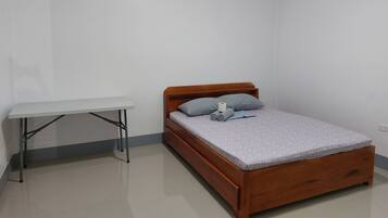 Quarto pequeno e bem organizado, com uma cama de madeira, uma escrivaninha branca e um travesseiro cinzento.