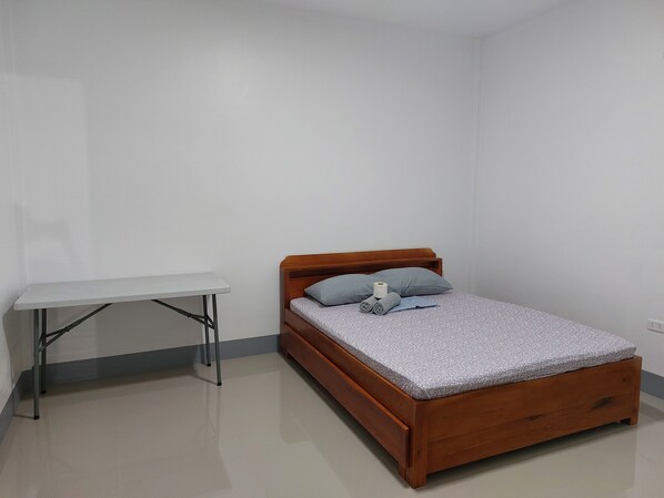 Quarto pequeno e bem organizado, com uma cama de madeira, uma escrivaninha branca e um travesseiro cinzento.