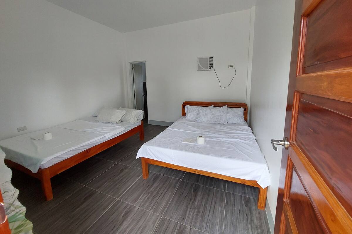 A room with two wooden beds, white bedding, a wooden door, a wall-mounted air conditioning unit, and a visible power outlet.