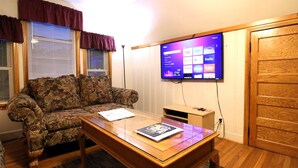 A living room with a floral sofa, a glass-top coffee table, and a flat-screen TV mounted on the wall displaying streaming services. - Cooperstown Baseball Rentals - Sky Box (Oneonta)
