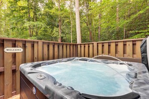 Ein Whirlpool, umgeben von einer Holzterrasse, mit einem 'WELCOME'-Schild, eingebettet in eine bewaldete Landschaft.