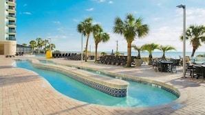 A swimming pool with a tiled edge, surrounded by lounge chairs and palm trees, with a building in the background.