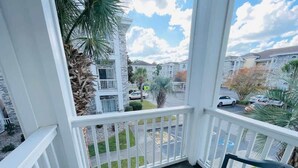 Ein überdachter Balkon mit weißen Geländern, einem schwarzen Stuhl und Blick auf ein Wohngebiet mit Häusern und einer Straße.