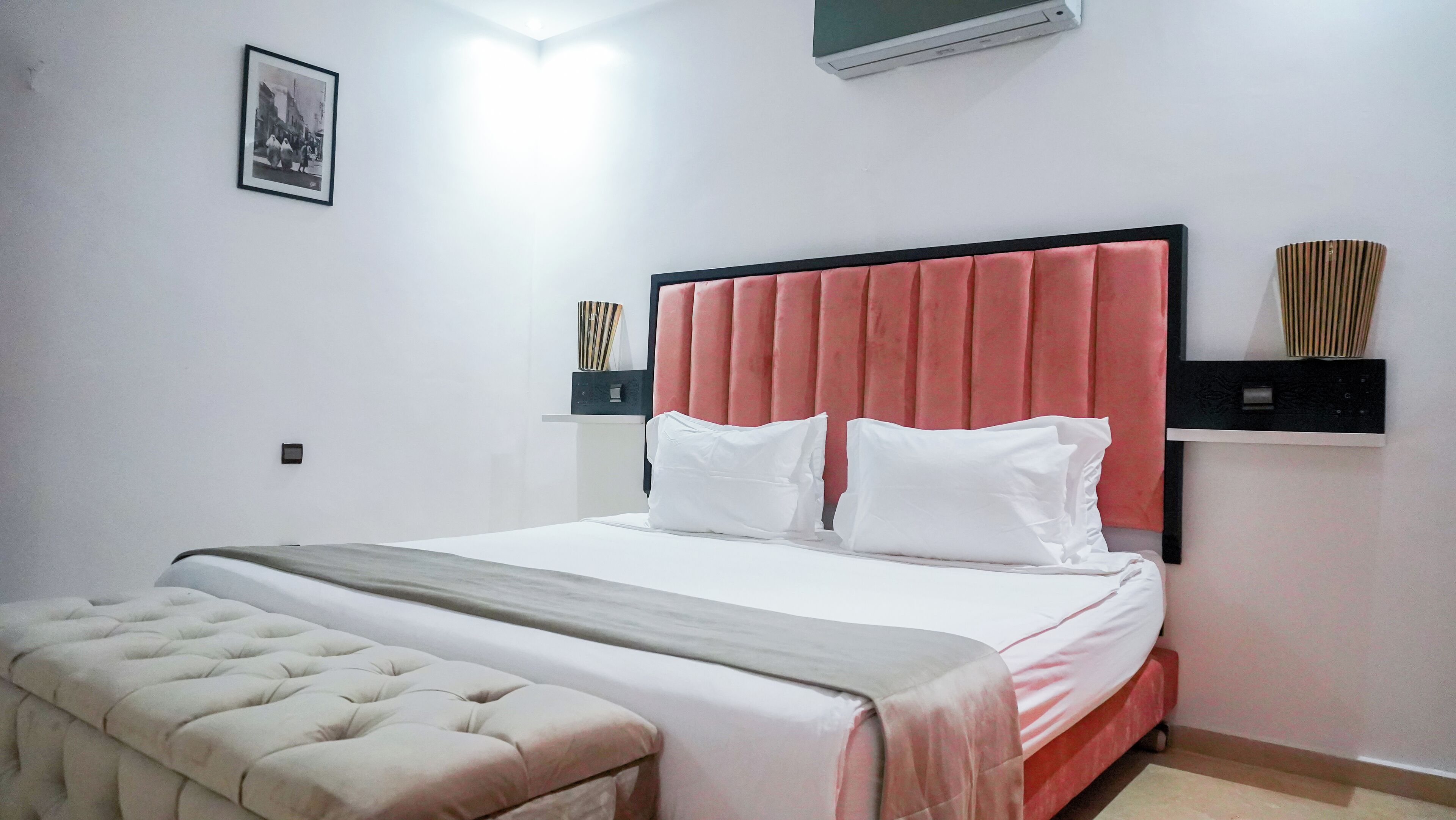 A neatly arranged bedroom with a bed, a bedside table, a wall-mounted air conditioner, and a framed picture on the wall.