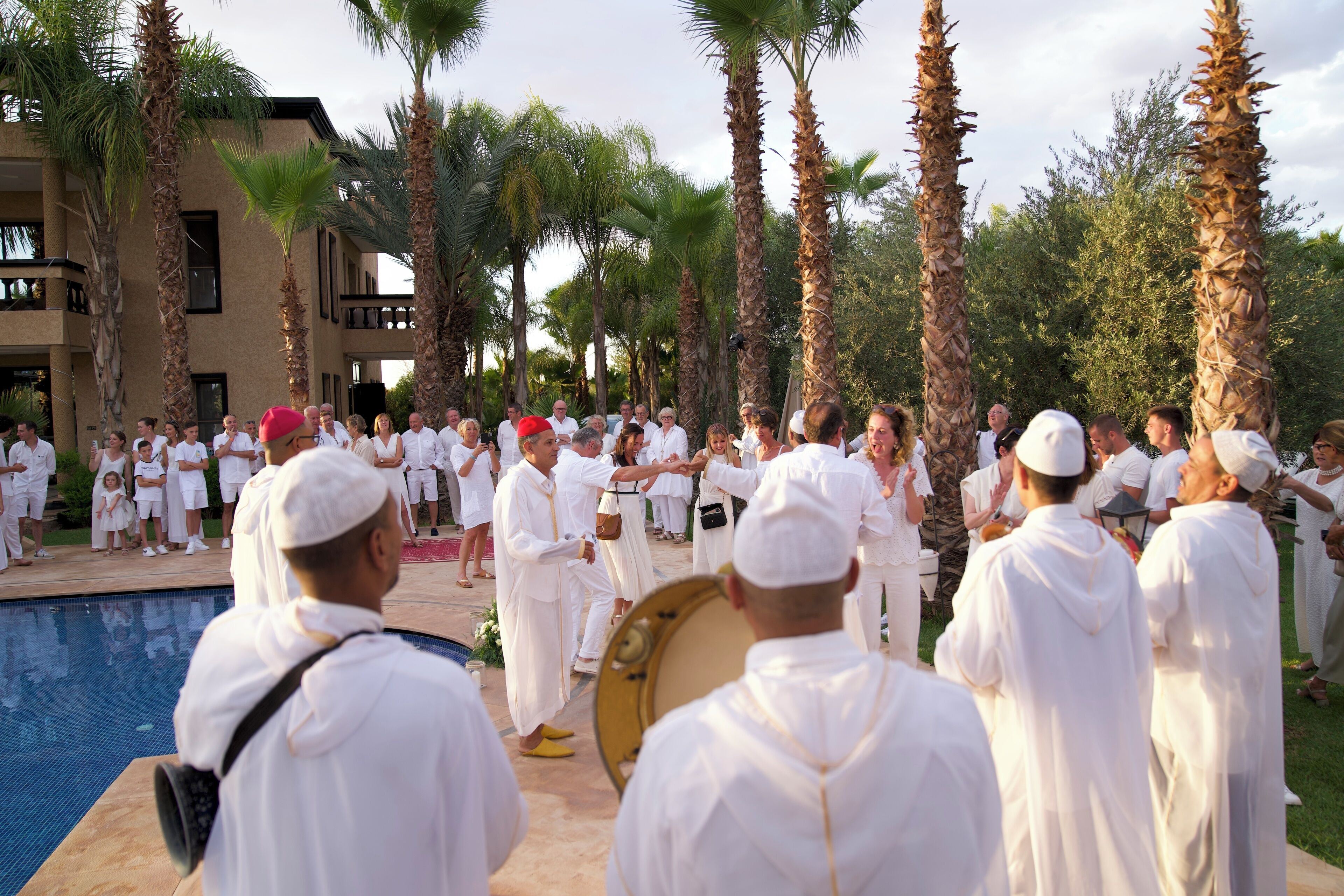 A group of people in white robes gathered around a pool area with palm trees and a building in the background.