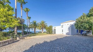 Une allée pavée mène à un bâtiment blanc doté d’une porte bleue, entouré de palmiers et surplombé d’un ciel d’un bleu éclatant.