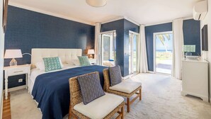 Une chambre avec un grand lit, deux fauteuils en osier, une table de chevet avec une lampe, et une vue sur l’extérieur grâce à une porte coulissante.