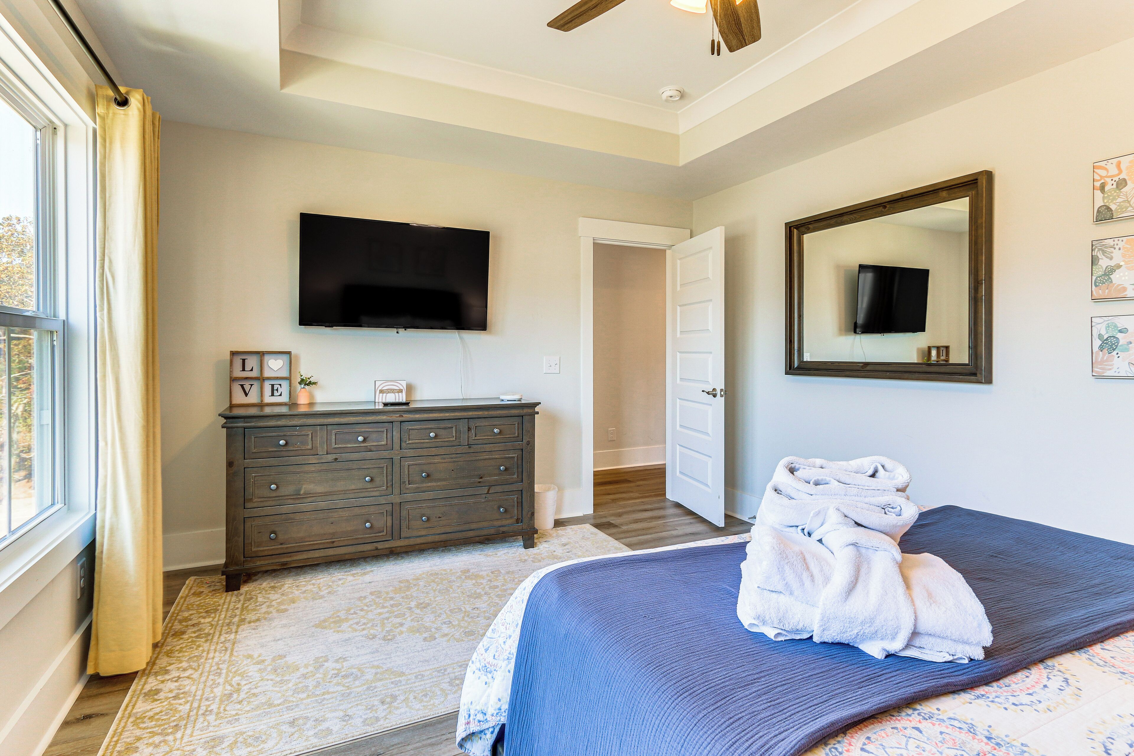 A bedroom with a bed, a dresser, a flat-screen TV, a mirror, and a window with curtains.