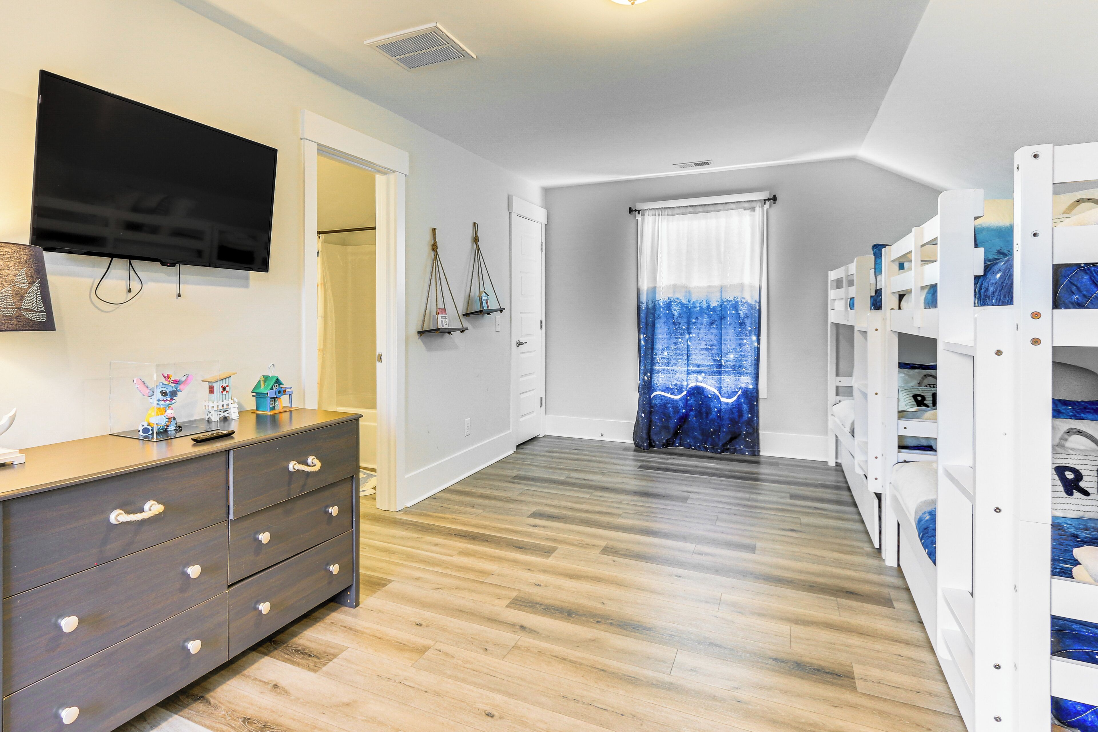 A room with a wooden floor, a TV mounted on the wall, a dresser with drawers, bunk beds, and a door leading to another room.