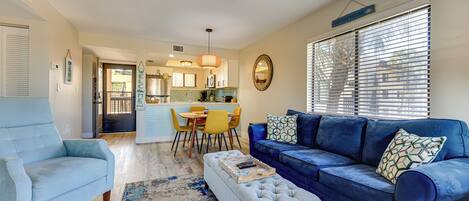 Uma sala de estar com um sofá azul, uma poltrona azul clara e um banquinho branco. É visível uma cozinha ao fundo, com uma mesa de jantar e cadeiras.