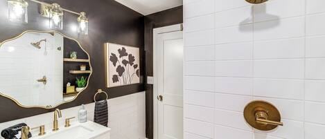 Une salle de bain avec un lavabo blanc, des robinets dorés, un miroir et une douche dotée d’un pommeau de douche doré.