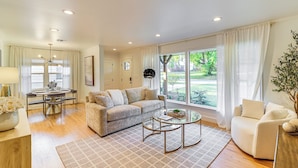 A spacious living room with a beige sofa, a glass coffee table, and a view of a garden through large windows. - Tulsa Retreat w/ Backyard Oasis & Putting Green! (Tulsa)