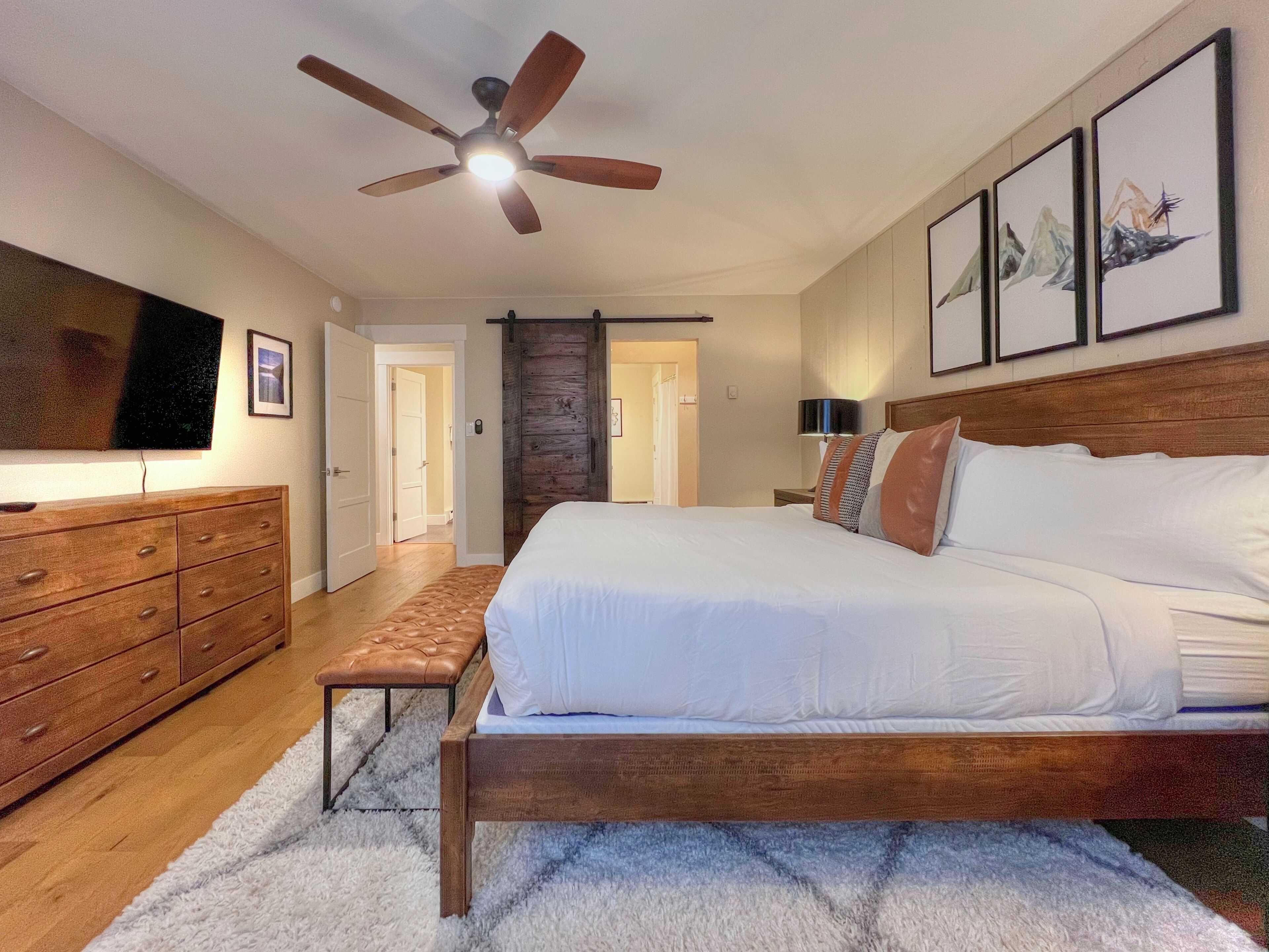 Une chambre à coucher avec un lit en bois, un ventilateur de plafond, une télévision, une commode et des œuvres d’art encadrées accrochées au mur.