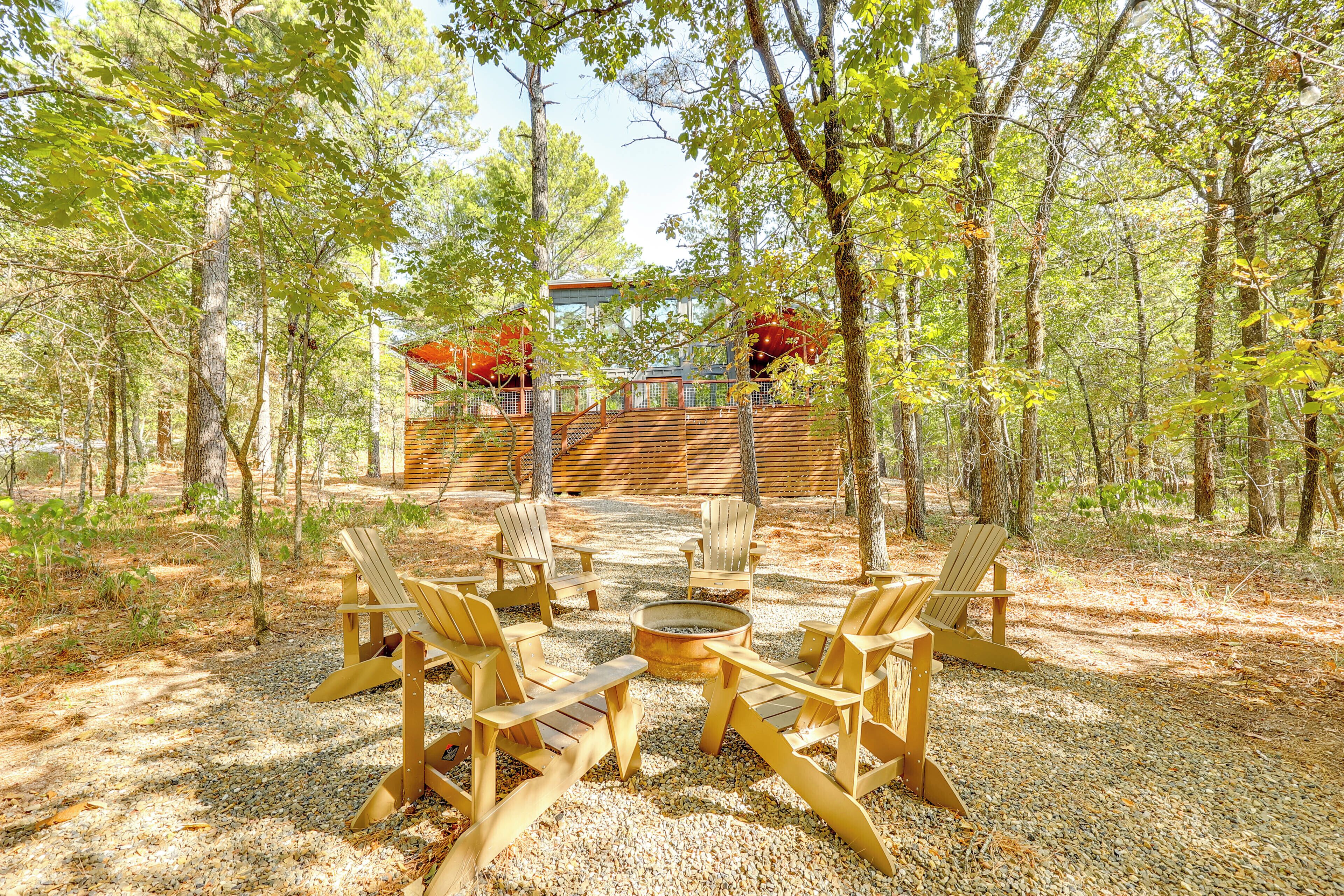 A forested area with a wooden deck, Adirondack chairs, and a fire pit.