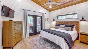 A bedroom with a wooden ceiling, a bed with multiple pillows, a nightstand, a dresser, and a window with a view of the outdoors. - Cozy Secluded Cabin in the Heart of Hochatown (Broken Bow)