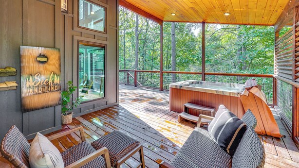 A wooden deck with outdoor seating, a hot tub, and a large window overlooking a forest. - Cozy Secluded Cabin in the Heart of Hochatown (Broken Bow)