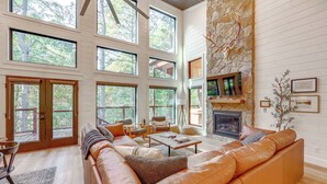 A spacious living room with a large leather sofa, a fireplace, and multiple windows offering views of the outdoors. - Cozy Secluded Cabin in the Heart of Hochatown (Broken Bow)