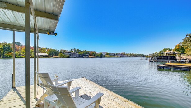 A wooden dock with two Adirondack chairs, a covered area, and a view of a calm lake with buildings in the background.