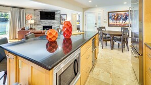 A modern kitchen with a central island, a dining area with a wooden table and chairs, and a living room with a sofa and a TV mounted on the wall.