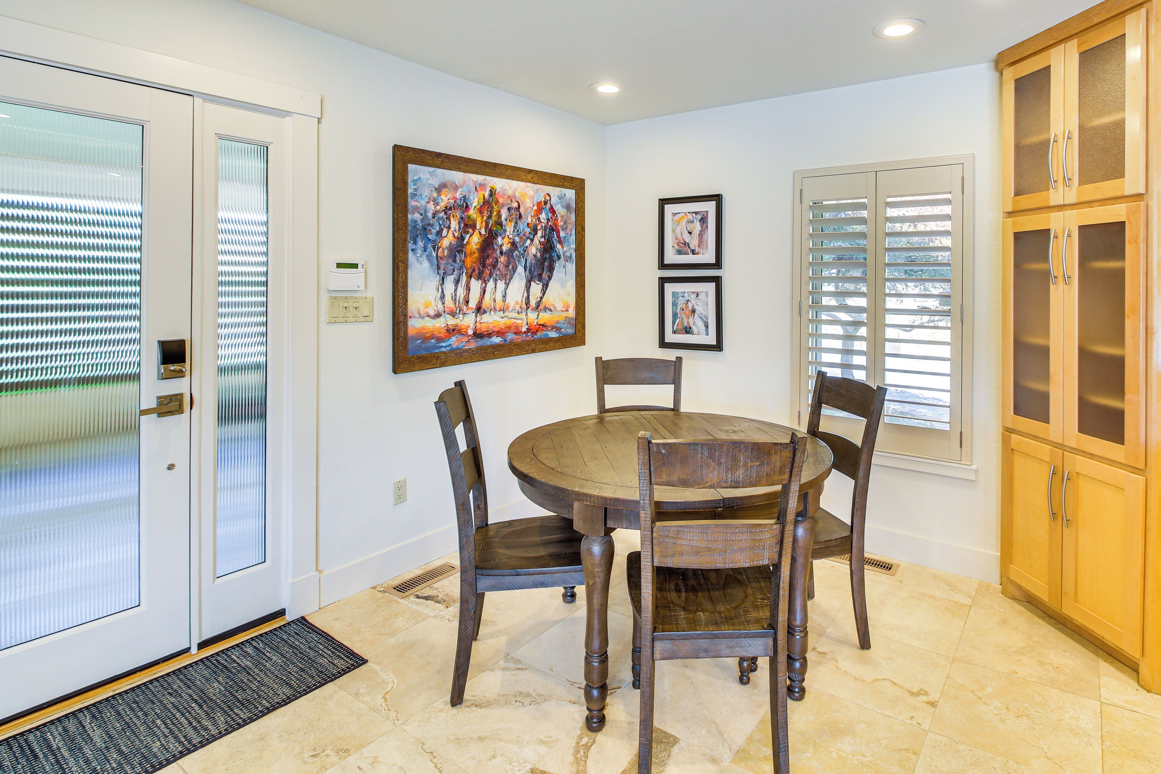 A dining area with a round wooden table and chairs, a painting on the wall, and a window with blinds.
