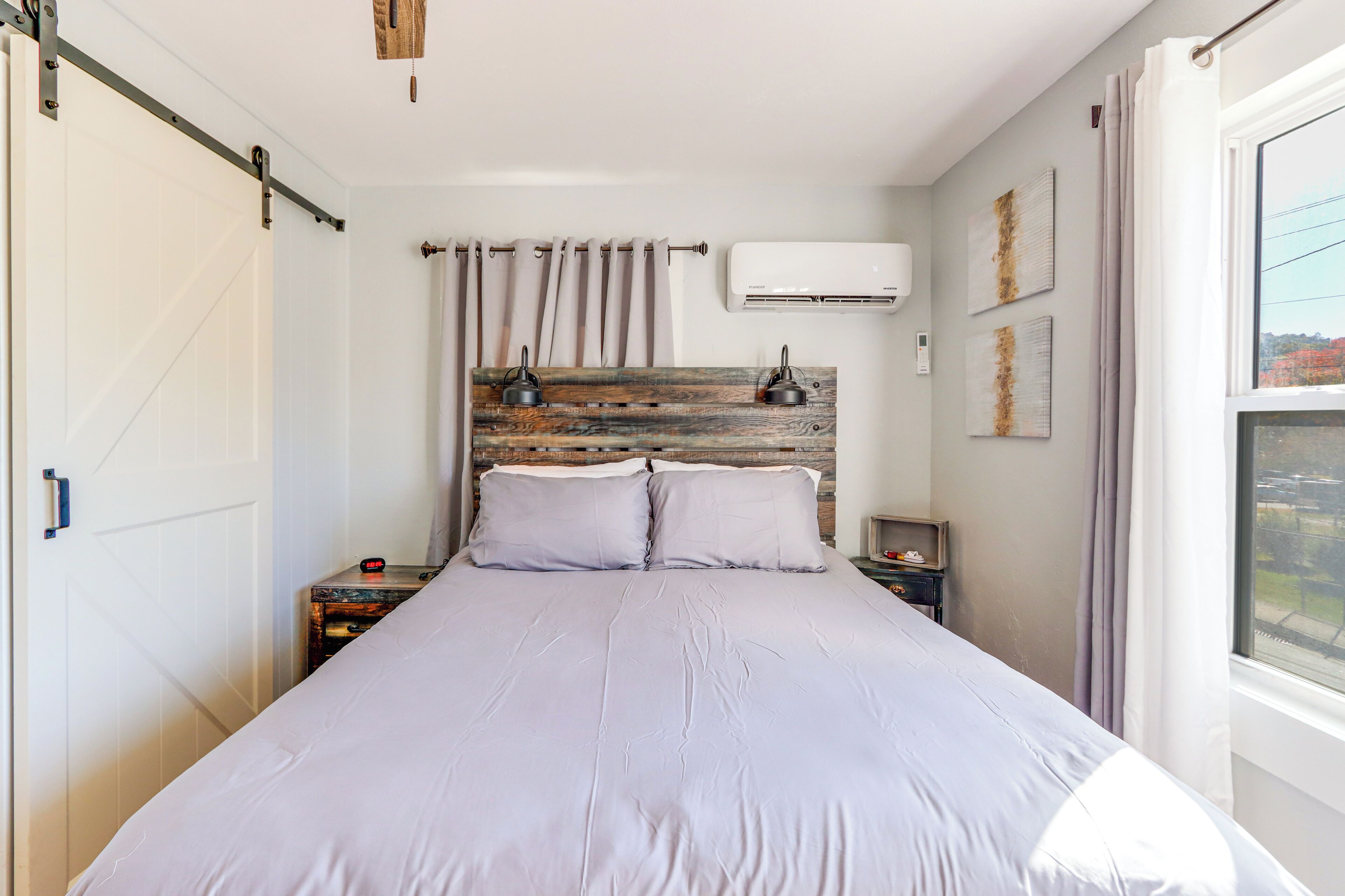 A bedroom with a bed, a headboard with a rustic design, an air conditioning unit, a nightstand, and a window with curtains.