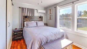 Une chambre à coucher comprenant un lit, une tête de lit en bois, un ventilateur de plafond, une table de chevet, un banc et une grande fenêtre donnant sur un beau paysage.