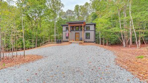 Une maison moderne à deux étages, dotée d’une allée en gravier et d’une terrasse en bois, et entourée d’une forêt.