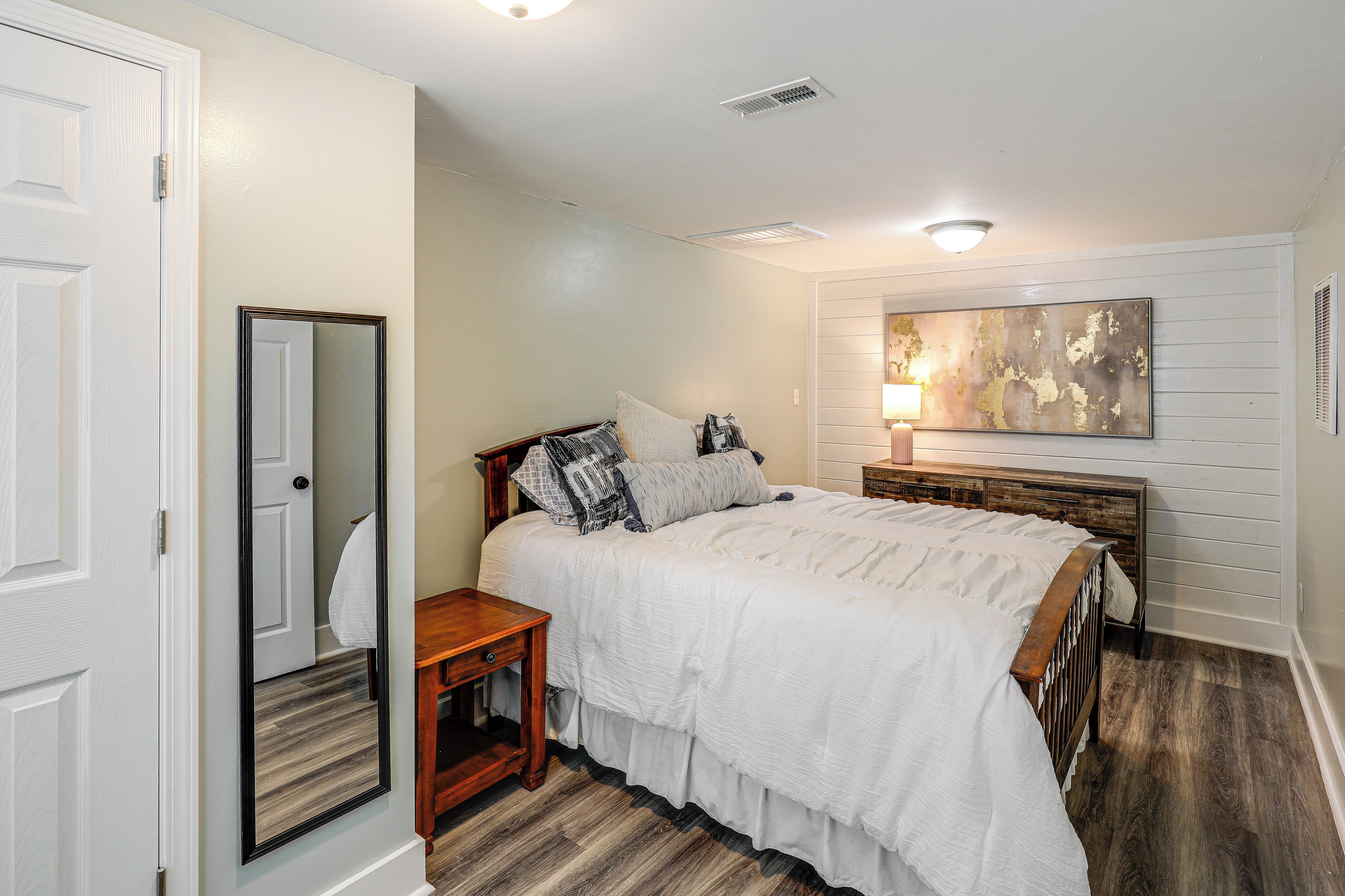 A bedroom with a bed, a nightstand, a mirror, a wall-mounted artwork, and a door.
