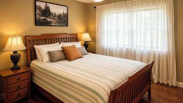 Une chambre avec un lit en bois, deux tables de chevet avec des lampes, une grande fenêtre avec des rideaux légers et un tableau encadré au mur.