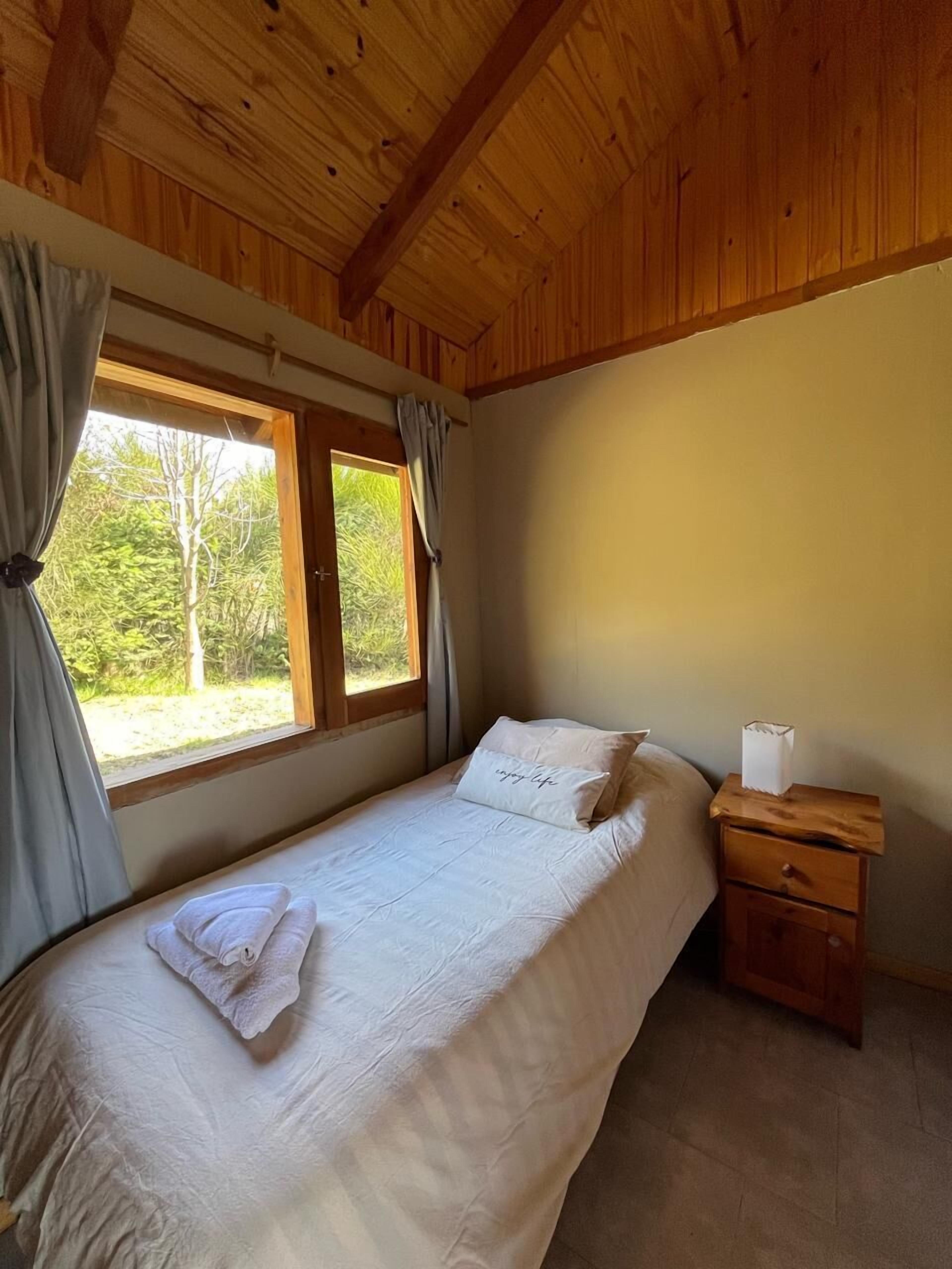A bedroom with a wooden ceiling, a bed with white linens, a nightstand with a tissue box, and a window with curtains.