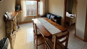 A wooden dining area with a table and chairs, a sofa, a window with curtains, and a bed in the adjacent room. - Cabañas Rayen  (Villa Traful)