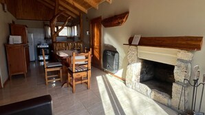 A cozy interior with a stone fireplace, wooden ceiling, and a dining area with chairs and a table. - Cabañas Rayen  (Villa Traful)