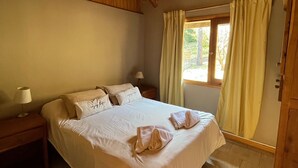 A bedroom with a wooden ceiling, a bed with white linens, two bedside tables with lamps, a window with curtains, and a door. - Cabañas Rayen  (Villa Traful)