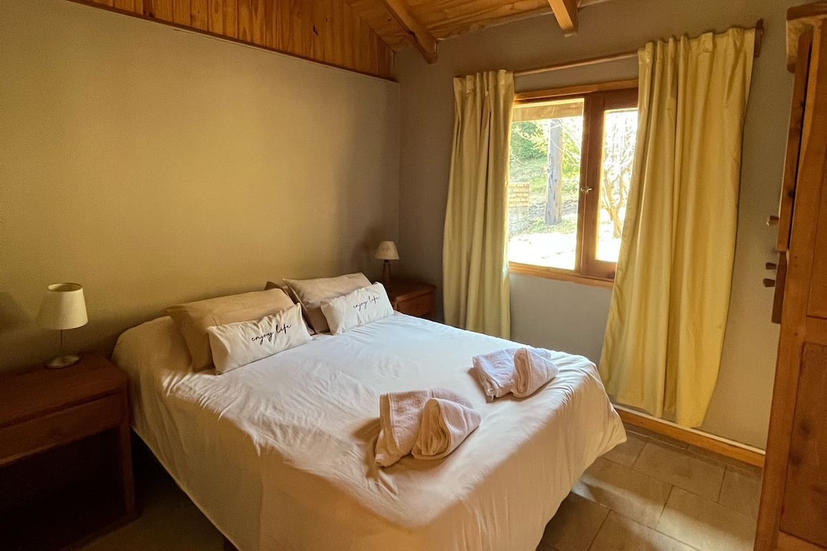 Un dormitorio con techo de madera, una cama con ropa de cama blanca, dos mesitas de noche con lámparas, una ventana con cortinas y una puerta.