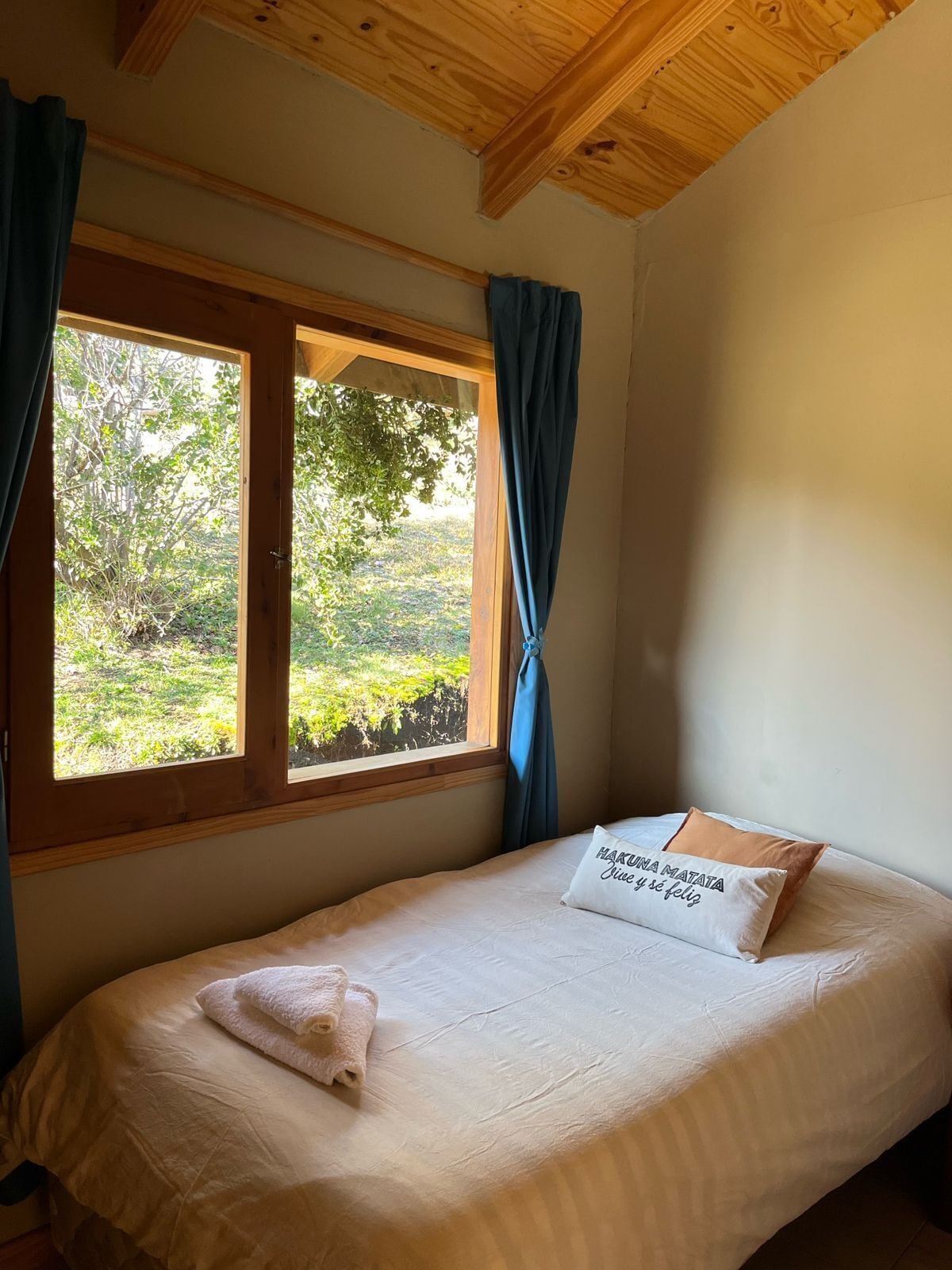 Una cama individual con colchón blanco y almohada con texto, una ventana con cortinas azules y vista a zonas verdes al exterior.